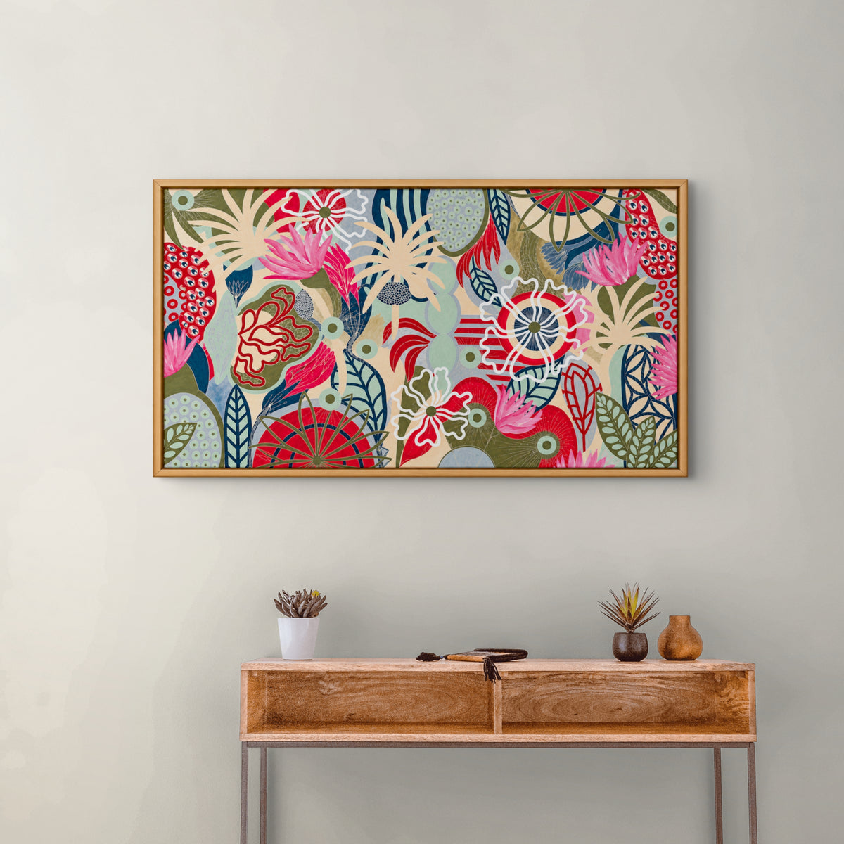 A framed abstract floral art print with a colorful pattern of pink, red, green, blue, and cream motifs, hanging on a light gray wall above a wooden console table with small decorative vases.