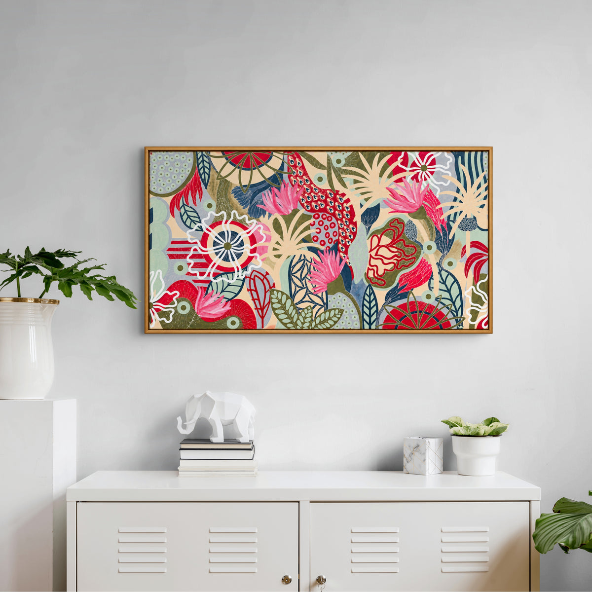 A framed rectangular wall art print with a colorful floral and botanical pattern hangs on a light gray wall above a white cabinet, styled with small plants and decorative objects in a bright interior setting.